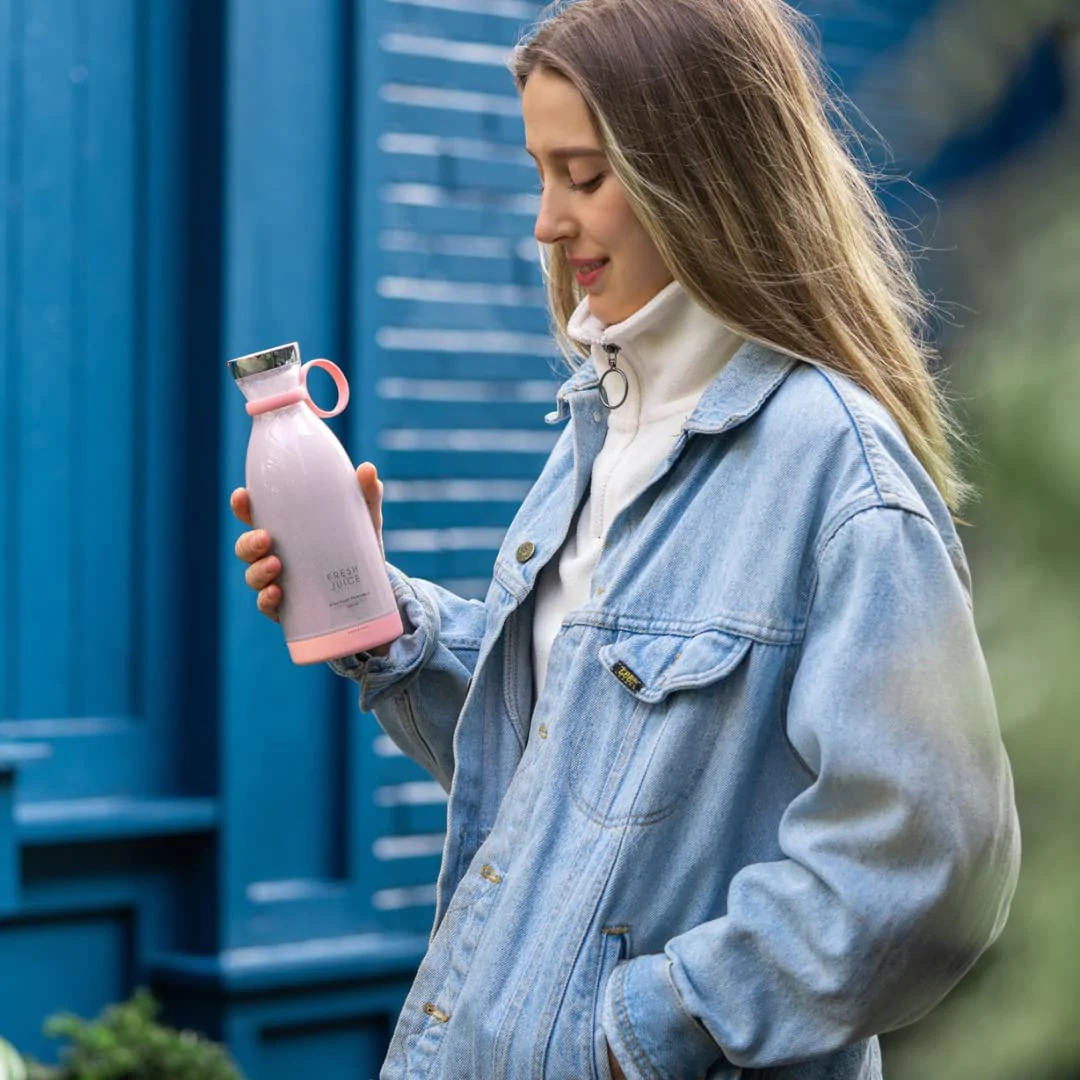 Eine lächelnde Frau hält einen rosa Fresh Juice Mixer und betrachtet einen frisch zubereiteten lila Smoothie.