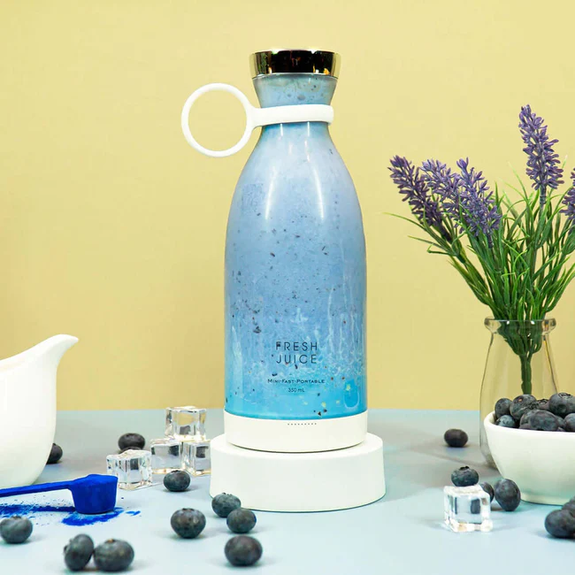 A white FreshJuice portable blender filled with blue smoothie, surrounded by blueberries and a plant.