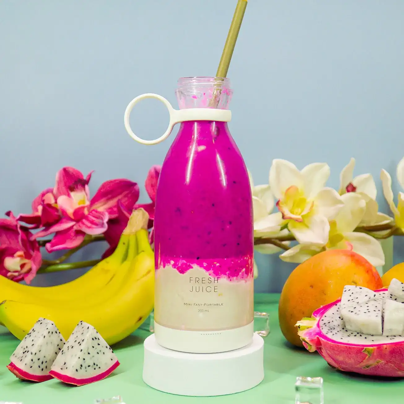 A white Fresh Juice blender filled with dark purple smoothie, surrounded by bananas, flowers, and dragon fruit.