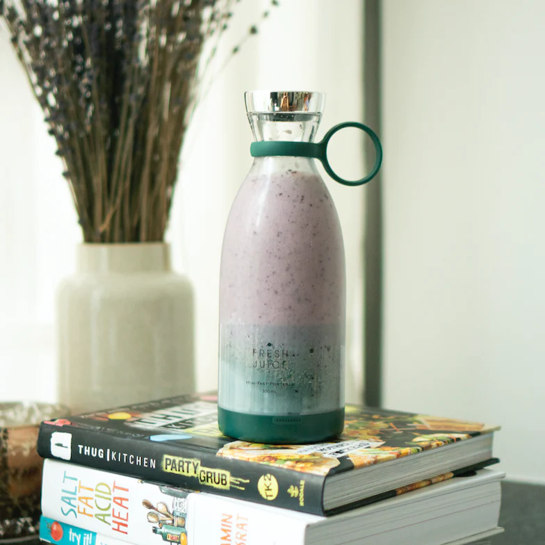 A green Fresh Juice blender, filled with a pink and white smoothie, sitting on two books in front of a plant in a vase.