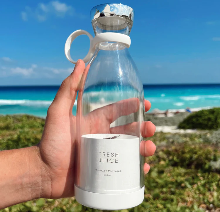 The Fresh Juice portable electric blender in front of the sea with a hand holding it to show its portability..